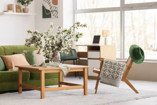 Yeşil kanepesi, koltuğu ve vazoda çiçekli dalları olan aydınlık oturma odasının içinde.