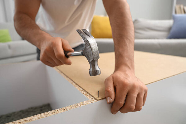 Young man with hammer assembling shelf unit at home, closeup