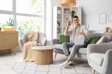 Ağırlık tartısı olan genç adam evdeki kanepede rahat görünüyor.