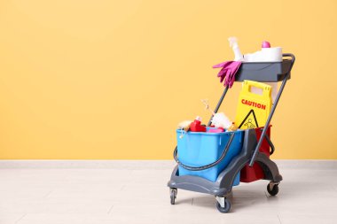 Temizlik malzemeleri ve sarı duvarın yanında uyarı levhası olan kapıcı vagonu.