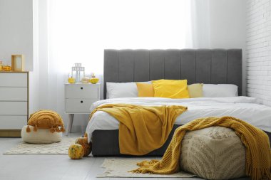 Büyük bir yatak, komodin, çekmeceler ve yatak odasının içinde sonbahar dekoru olan puf.