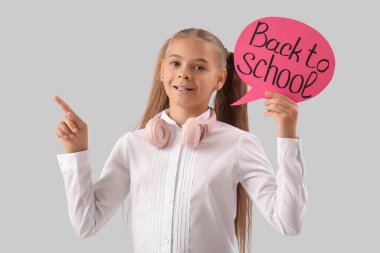 Okul üniformalı, elinde konuşma balonuyla okula geri dönen ve gri arkaplandaki bir şeyi işaret eden sevimli küçük bir kız.