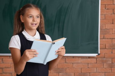 Okul üniformalı şirin bir kız sınıfın yanında yazı tahtası okuyor.