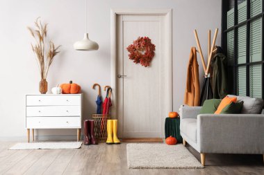 Koridordaki beyaz duvarın yanında sonbahar balkabaklı, şemsiyeli ve çizmeli kapı paspası.