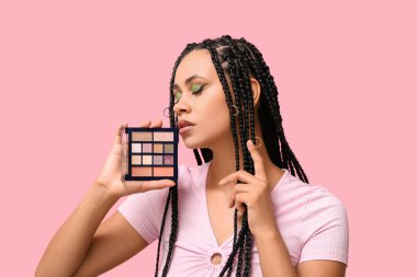 Saç örgüleri ve güzel makyajlı genç Afro-Amerikalı kadın pembe arka planda bir palet göz falı tutuyor.