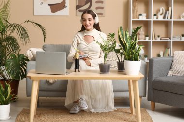 Kulaklık takan, bitkilerle birlikte evde podcast yapan genç bir kadın.