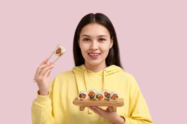 Güzel bir Asyalı kadın elinde çubuklar ve nefis suşili ahşap tepsi soluk pembe arka planda dürüm tutuyor.