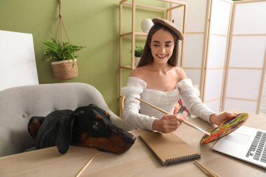 Sevimli Doberman köpeği ve evde elinde boya paleti olan bereli güzel ve mutlu bir kadın.