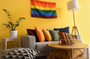 Oturma odasının içinde kanepe, masa ve sarı duvarda LGBT bayrağı var.