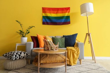 Oturma odasının içinde kanepe, masa ve sarı duvarda LGBT bayrağı var.