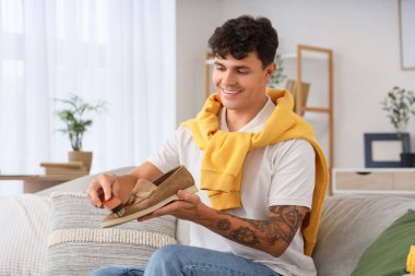 Genç adam evdeki kanepede fırçayla süet ayakkabı temizliyor.