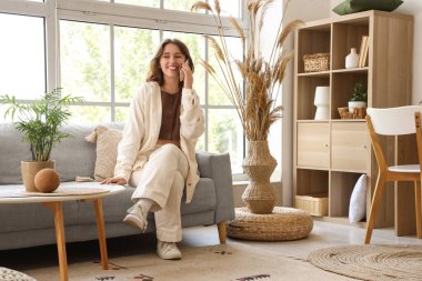 Oturma odasındaki koltukta cep telefonuyla konuşan genç bir kadın.