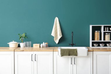 Mutfaktaki koyu mavi duvarın yanındaki ahşap tezgahın üzerinde beyaz seramik lavabo ve raf ünitesi.