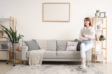 Dizüstü bilgisayarı olan genç bir kadın oturma odasında gri koltukta oturuyor.