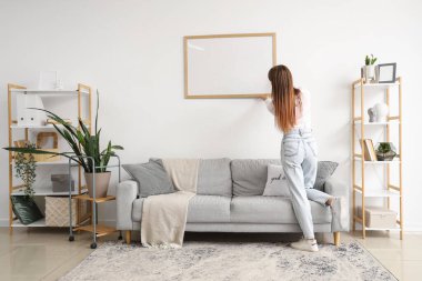 Genç bir kadın oturma odasındaki ışık duvarına boş bir çerçeve asıyor.