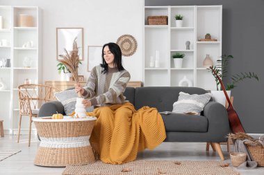 Evdeki kanepede sıcak ekose ve vazoyla oturan güzel, genç ve mutlu bir kadın.