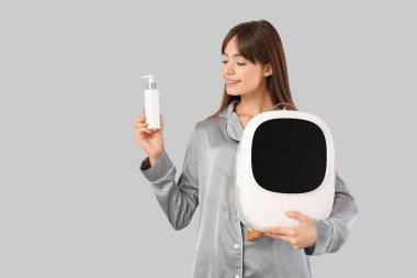 Gri arka planda kozmetik ürünleri için küçük buzdolabı olan güzel genç bir kadın.