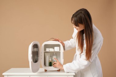 Bej arka planda kozmetik ürünleri için küçük buzdolabı olan güzel genç bir kadın.