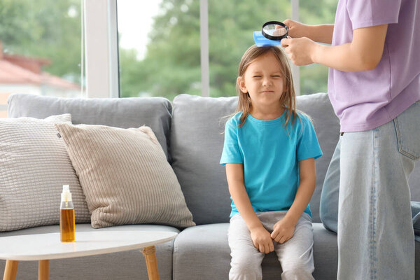 Mother with magnifier combing her little daughter's hair with pediculosis at home