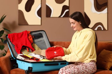 Evdeki kanepede bavulu ve ilk yardım çantasıyla oturan mutlu genç kadın. Seyahat kavramı