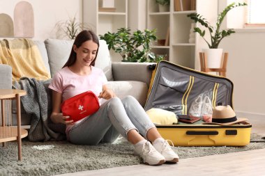 Evinde yerde oturan bavulu ve ilk yardım çantasıyla mutlu genç bir kadın. Seyahat kavramı