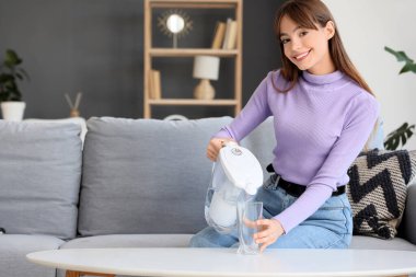 Evdeki masada filtre testisinden bardağa su döken genç bir kadın.