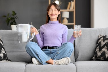 Su filtresi ve cam sürahi ile evdeki kanepede meditasyon yapan genç bir kadın.