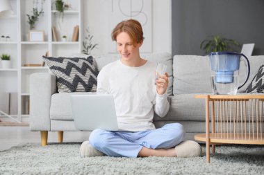 Evde dizüstü bilgisayar kullanan cam ve su filtresi olan genç bir adam.