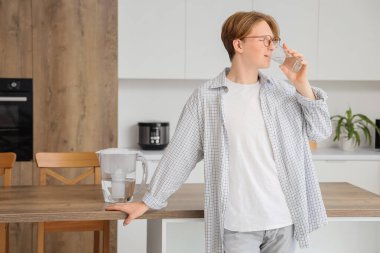 Genç adam mutfaktaki masada filtre sürahi ile su içiyor.