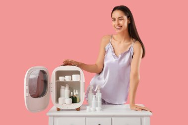 Küçük kozmetik buzdolabını açan mutlu genç kadın pembe arka planda.