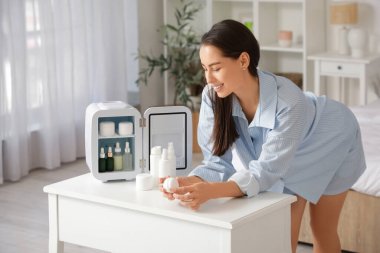 Evinde küçük kozmetik buzdolabı olan, masanın yanındaki mutlu genç kadın.