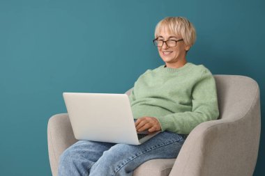 Dizüstü bilgisayarı olan yaşlı kadın mavi duvarın yanında oturuyor.