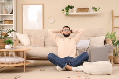 Evde dizüstü bilgisayarlı, gözlüklü genç sakallı bir adam oturuyor.