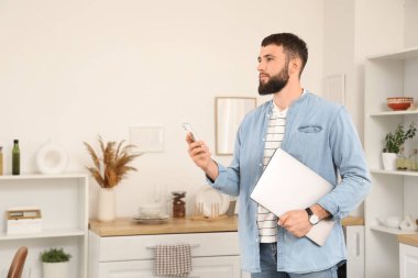 Genç sakallı adam mutfakta cep telefonu ve dizüstü bilgisayarla çalışıyor.