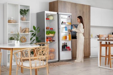 Genç bir kadın mutfaktaki temiz yiyeceklerle dolu açık buzdolabından su şişesini alıyor.