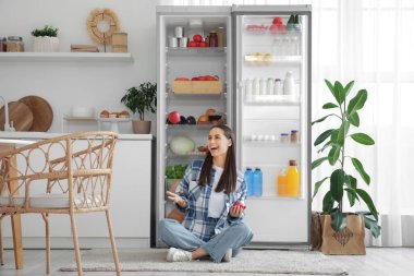 Genç bir kadın taze elmalarla mutfaktaki yiyecek dolu buzdolabının yanında oturuyor.