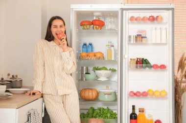 Genç bir kadın mutfaktaki açık buzdolabının yanında taze elma yiyor.