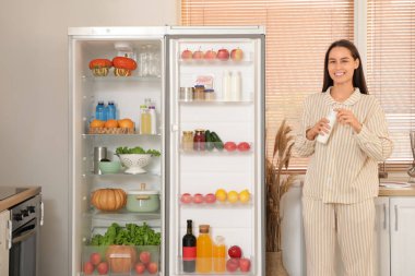 Mutfaktaki açık buzdolabının yanında yoğurt şişesi olan genç bir kadın.