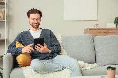 E-okuyuculu genç adam evdeki kanepede oturuyor.