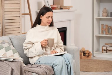 Evdeki kanepede bir fincan kakao içip kitap okuyan güzel bir kadın.