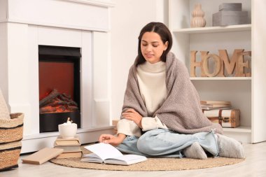 Evinde şöminenin yanında bir fincan kakao ve ekoseli kitap okuyan güzel, mutlu bir kadın.