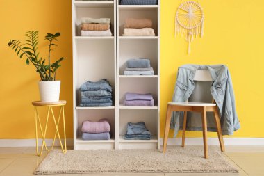 Katlanmış elbiseli raf ünitesi, sandalye ve çiçek renkli duvarın yanında.