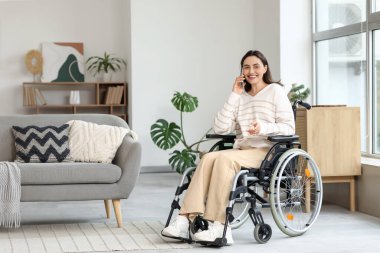 Tekerlekli sandalyedeki genç kadın evde cep telefonuyla konuşuyor.