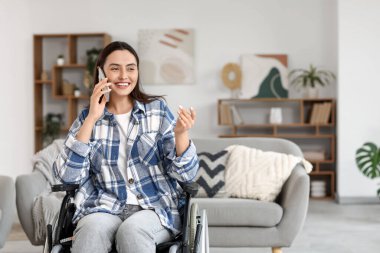 Tekerlekli sandalyedeki genç kadın evde cep telefonuyla konuşuyor.