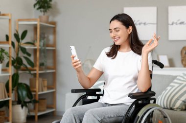 Tekerlekli sandalyedeki genç kadın evde cep telefonu kullanıyor.
