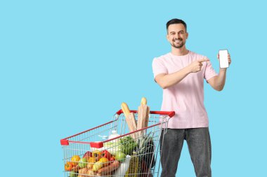 Mavi arka planda cep telefonunu gösteren alışveriş arabalı mutlu genç adam.