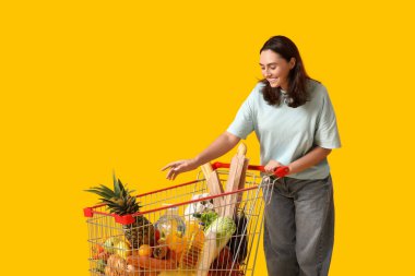 Sarı arka planda alışveriş arabası olan güzel, genç ve mutlu bir kadın.