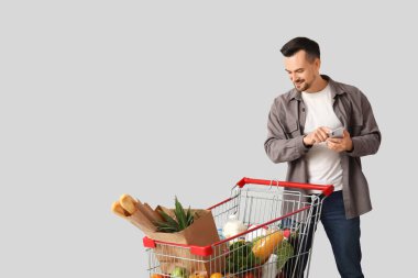 Gri arka planda tam alışveriş arabası ve cep telefonu olan mutlu genç bir adam.