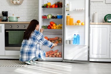 Genç bir kadın mutfaktaki açık buzdolabından taze havuç alıyor.