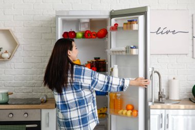 Genç Asyalı kadın mutfaktaki taze yiyeceklerle dolu açık buzdolabının yanında.